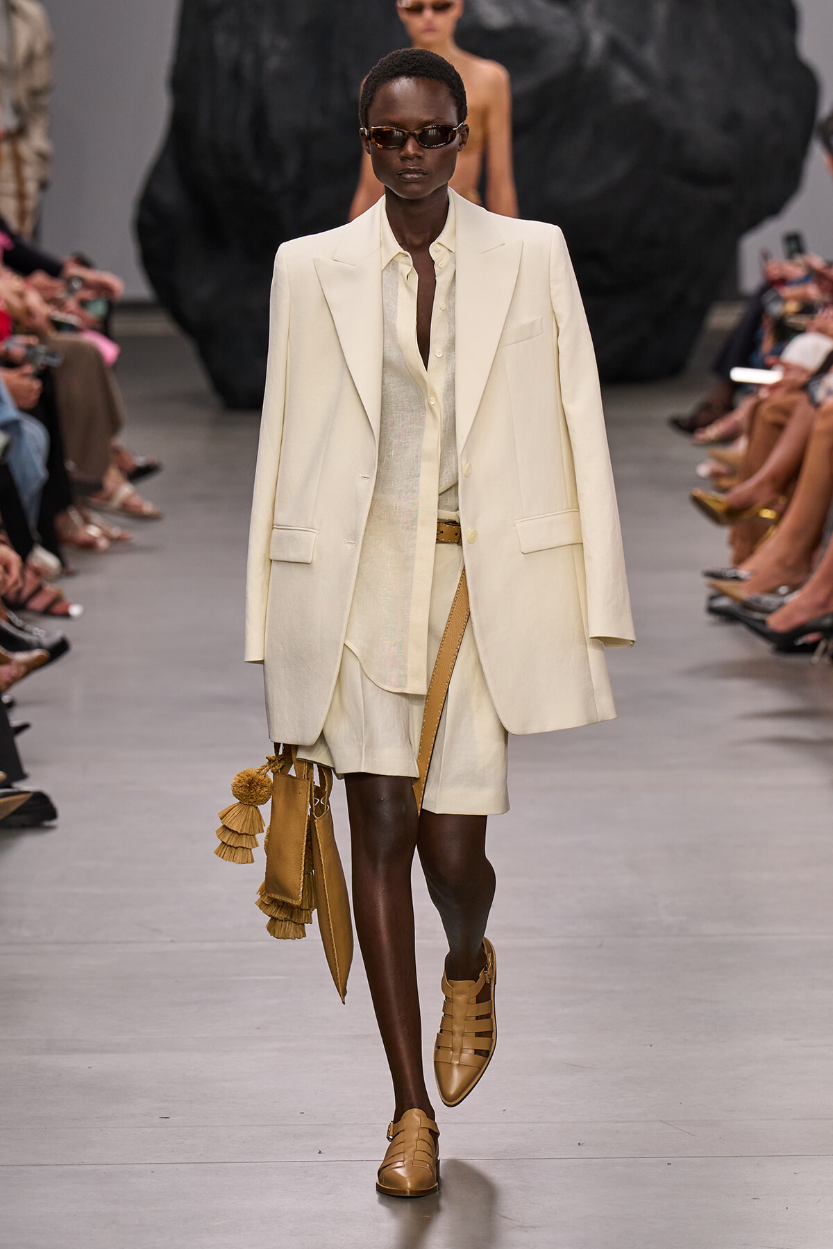 Model walking a fashion show in an oversized cream suit with shorts, tan sandals, and a tan bag with tassels.