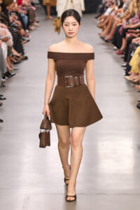 Model walks a brown off-shoulder top with wide belt and A-line mini skirt on a runway, carrying a brown handbag and wearing brown sandals.