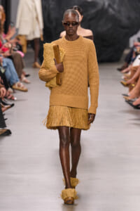 Model walking a runway in a camel knit sweater with a fringe mini skirt, sunglasses, and fringed sandals, carrying a textured clutch.