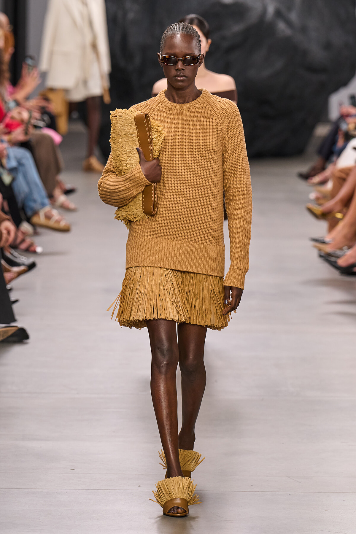 Model walking a runway in a camel knit sweater with a fringe mini skirt, sunglasses, and fringed sandals, carrying a textured clutch.