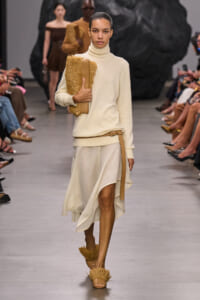 Model walks a runway in an ivory turtleneck sweater and flowing cream skirt, carrying a textured tan fringed clutch with a tan belt accessory.