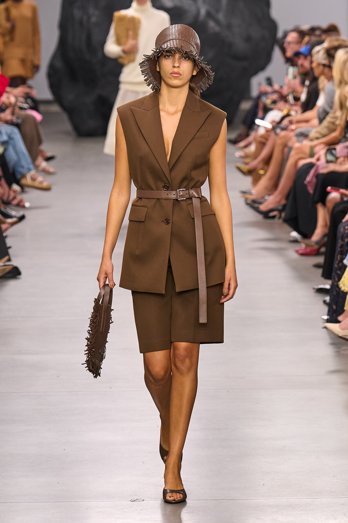 Model walks the runway in a brown sleeveless blazer and matching shorts with a belt, wearing a fringed brown hat and carrying a circular fringed handbag.