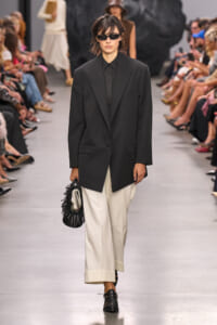 Model on a runway wearing an oversized black blazer and black shirt with white wide-leg pants, black loafers, sunglasses, and a black fringe handbag.
