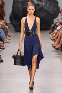 Model walks the runway in a navy blue plunging-V dress with a wide black belt, black heels, and a fringed black woven handbag.