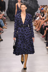Model walks the runway wearing a navy blue, knee‑length coat covered in black floral appliqués with a black belt and pointed black heels.