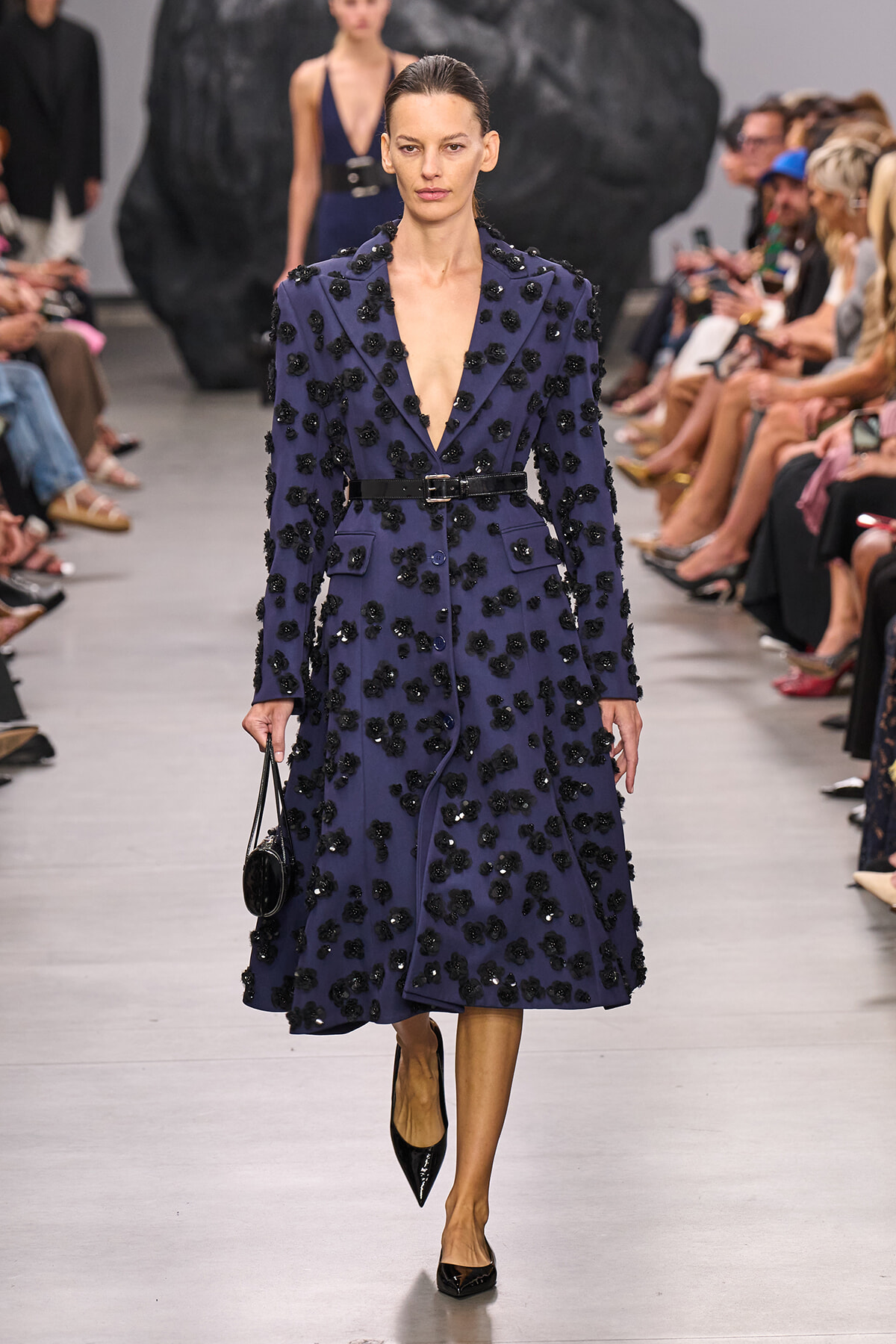 Model walks the runway wearing a navy blue, knee‑length coat covered in black floral appliqués with a black belt and pointed black heels.