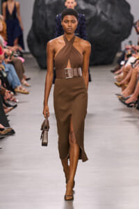 Model walks a fashion runway in a brown halter-wrap dress with a wide belt and thigh-high slit, carrying a small brown handbag.