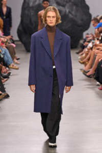 Male model walking a fashion runway in a purple overcoat over a brown turtleneck and black pants, audience on sides.