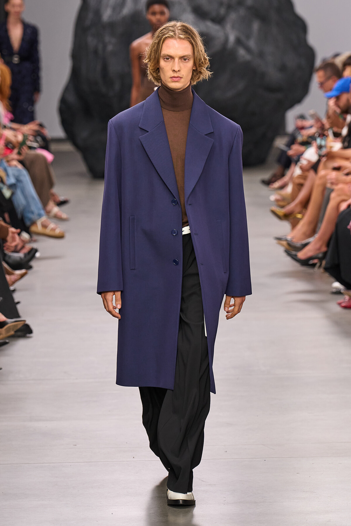 Male model walking a fashion runway in a purple overcoat over a brown turtleneck and black pants, audience on sides.