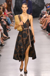 Model walks the runway in a black-and-brown floral halter dress with a brown belt, holding a textured beige clutch under her left arm, and wearing brown pointed-toe heels.