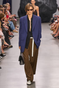 Model walks the runway in a cobalt blue blazer over a black shirt, brown wide-leg trousers, and a white belt, carrying a black handbag while wearing sunglasses.