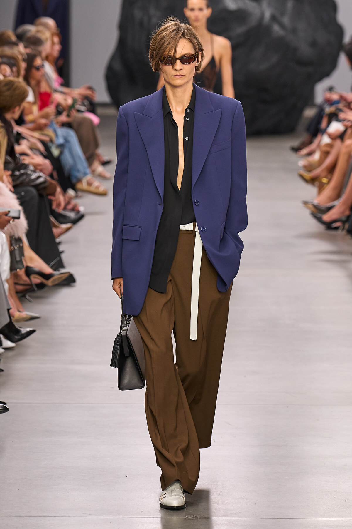 Model walks the runway in a cobalt blue blazer over a black shirt, brown wide-leg trousers, and a white belt, carrying a black handbag while wearing sunglasses.