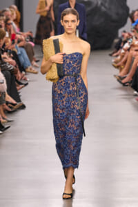 Model walking a runway in a blue lace strapless dress, holding a woven tan clutch.