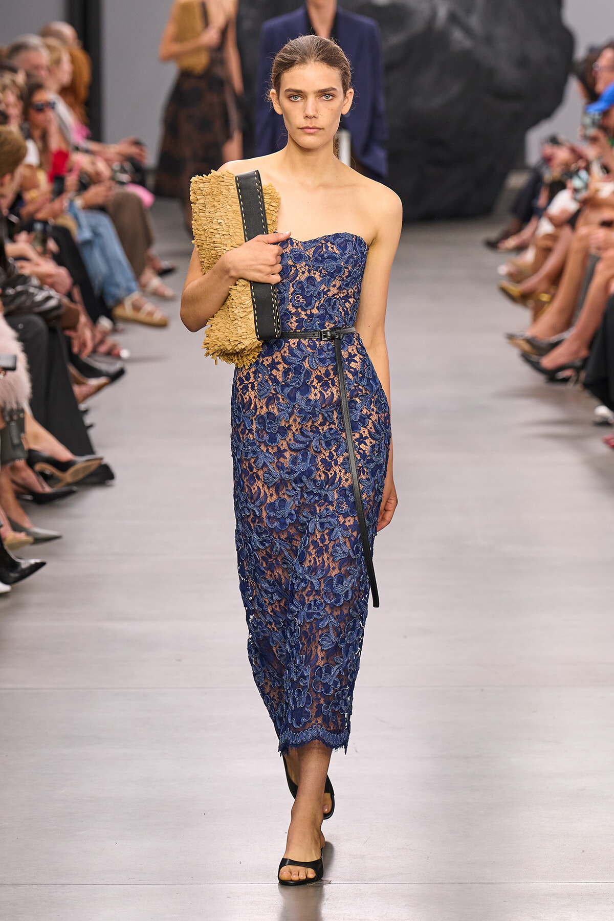 Model walking a runway in a blue lace strapless dress, holding a woven tan clutch.