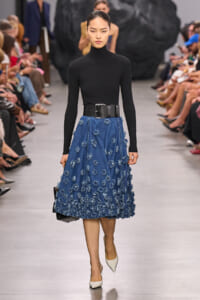 Model on a runway wearing a black long-sleeve turtleneck, a blue denim A-line skirt with scattered blue floral appliqués, a wide black belt, white pointed heels, and a small black handbag.