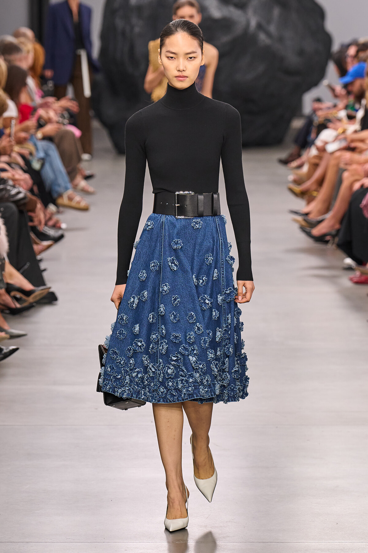 Model on a runway wearing a black long-sleeve turtleneck, a blue denim A-line skirt with scattered blue floral appliqués, a wide black belt, white pointed heels, and a small black handbag.
