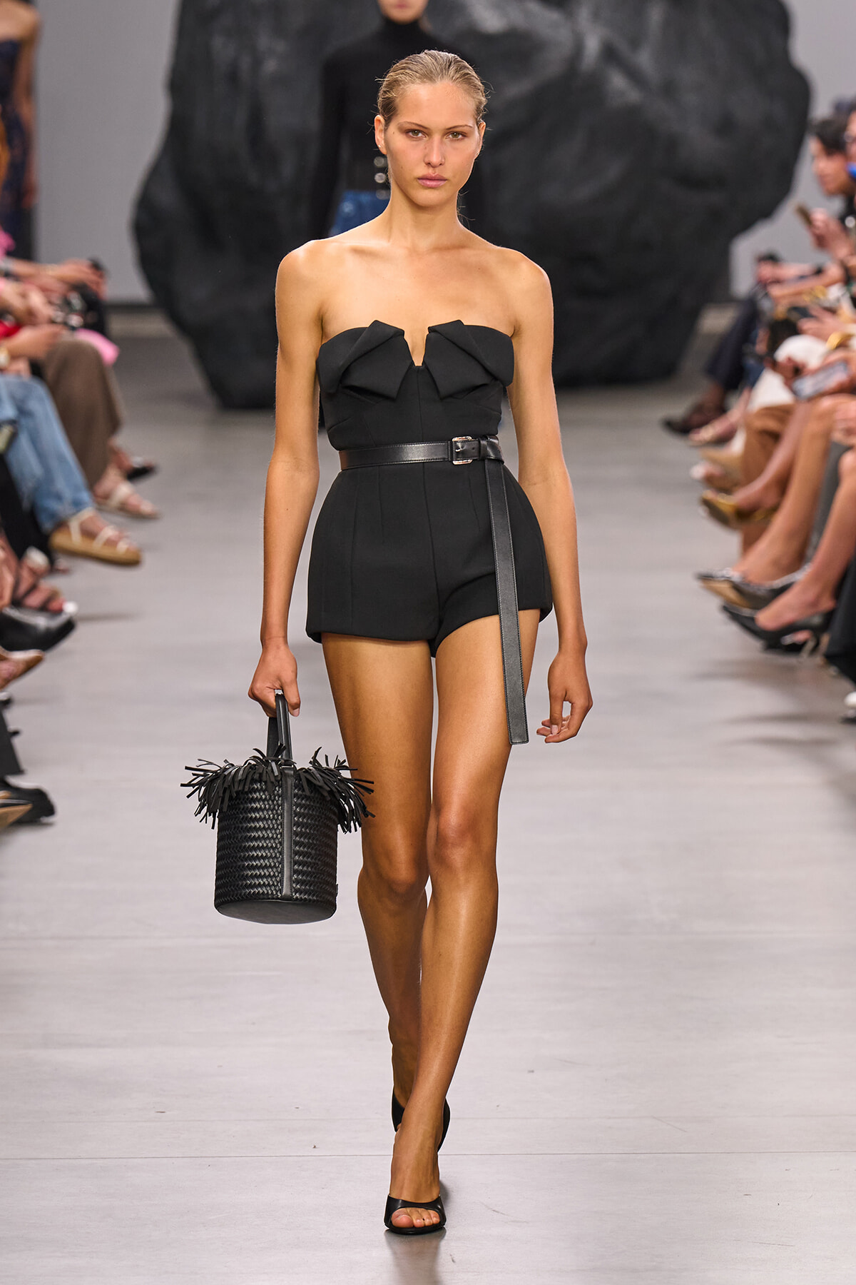 Model walking a black strapless romper with oversized bow, cinched waist, and a textured black bucket bag with fringe on a runway.