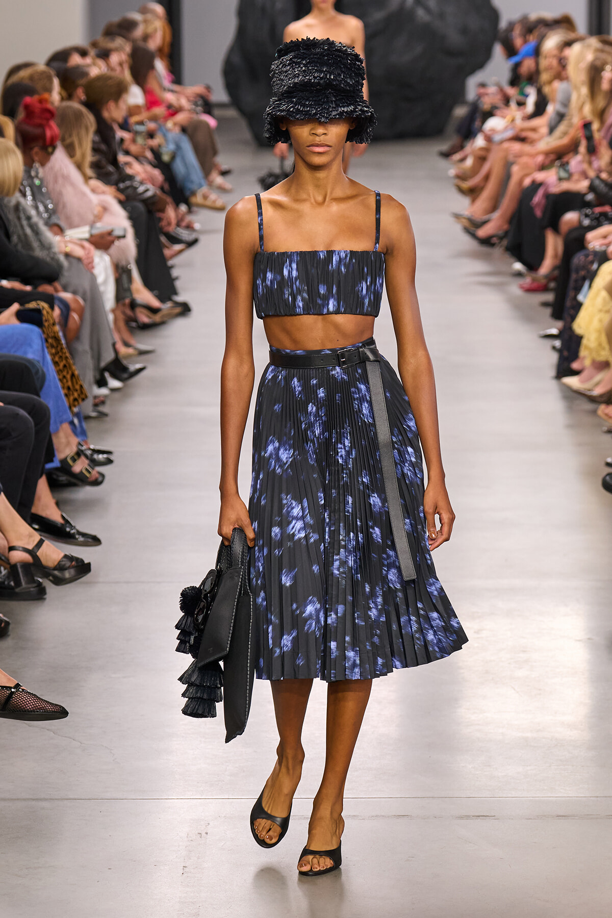 Model walks the runway in a navy tie-dye crop top with thin straps and a matching pleated midi skirt, cinched with a black belt, plus a black shaggy bucket hat and a black handbag with fringe.