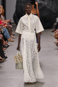 Model on a runway wearing a crisp white button-down top with wide sleeves and a long white lace skirt, belted, carrying a woven handbag.