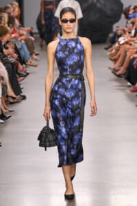 Model on a runway wearing a blue and black abstract print sleeveless halter-neck jumpsuit with a black belt, dark sunglasses, and a small black fringe handbag.