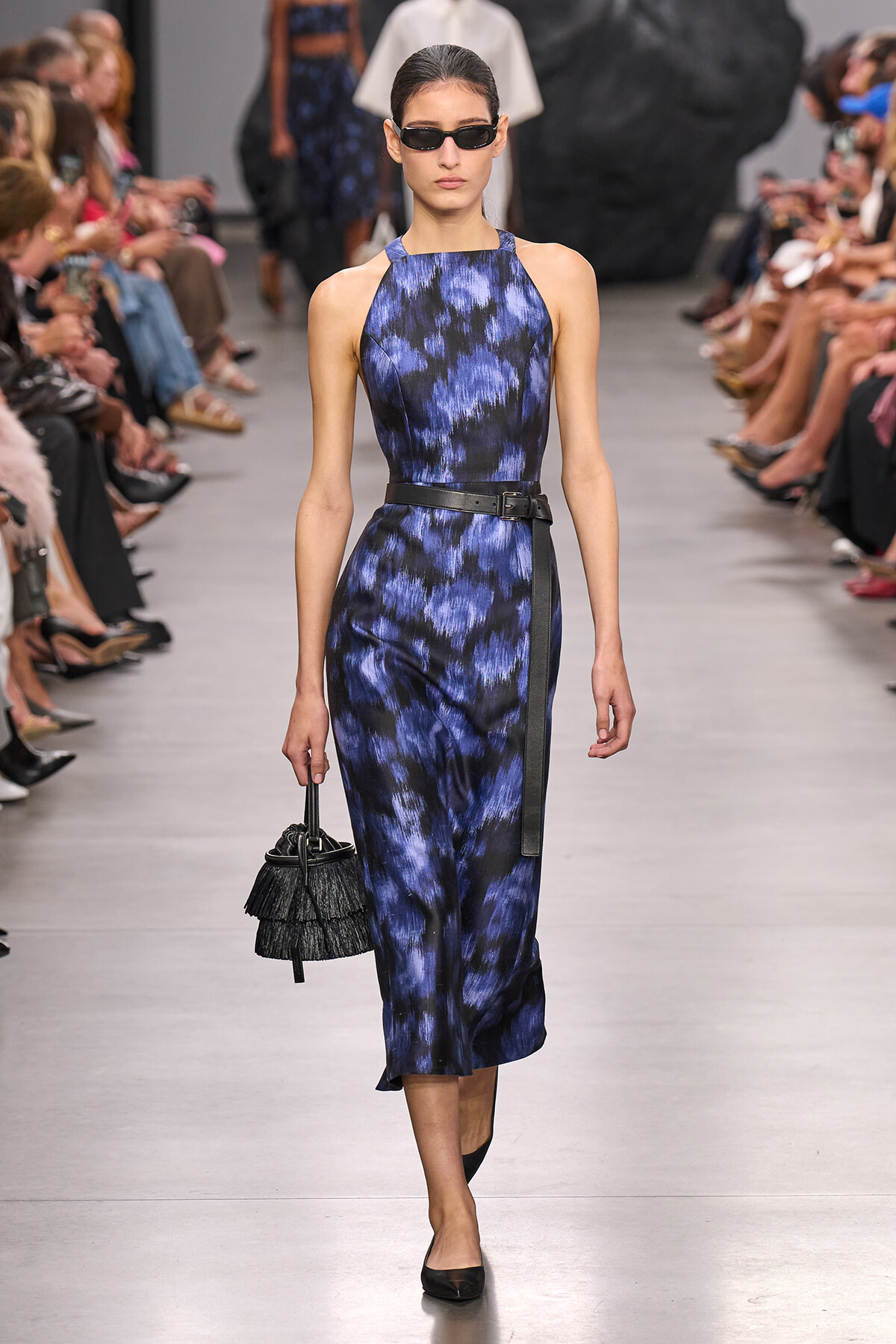 Model on a runway wearing a blue and black abstract print sleeveless halter-neck jumpsuit with a black belt, dark sunglasses, and a small black fringe handbag.