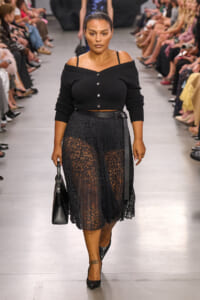 Model walking a runway in a black off-shoulder cardigan with a lace midi skirt, black belt, and pointed heels, carrying a structured black bag.
