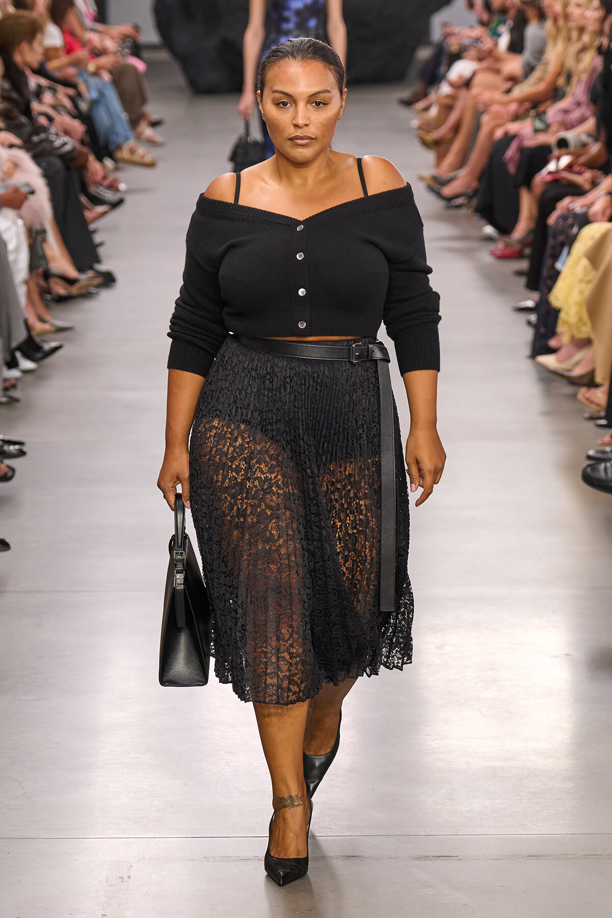 Model walking a runway in a black off-shoulder cardigan with a lace midi skirt, black belt, and pointed heels, carrying a structured black bag.