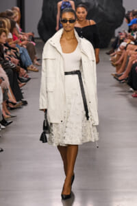 Model walking a fashion runway in an oversized white coat over a white top and textured white skirt, paired with a black belt and black heels, wearing dark sunglasses and carrying a small black handbag.