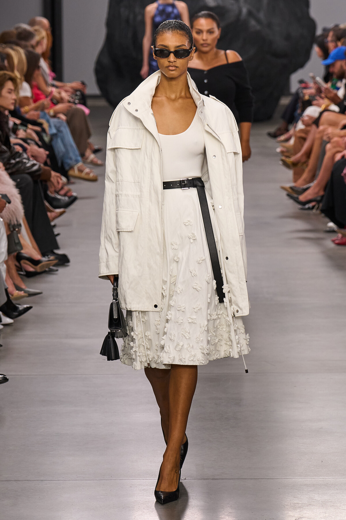 Model walking a fashion runway in an oversized white coat over a white top and textured white skirt, paired with a black belt and black heels, wearing dark sunglasses and carrying a small black handbag.