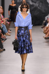 Model walking a runway in a light blue button-down blouse with rolled sleeves, a dark blue printed midi skirt, and a black belt, carrying a black fringe handbag while wearing sunglasses.