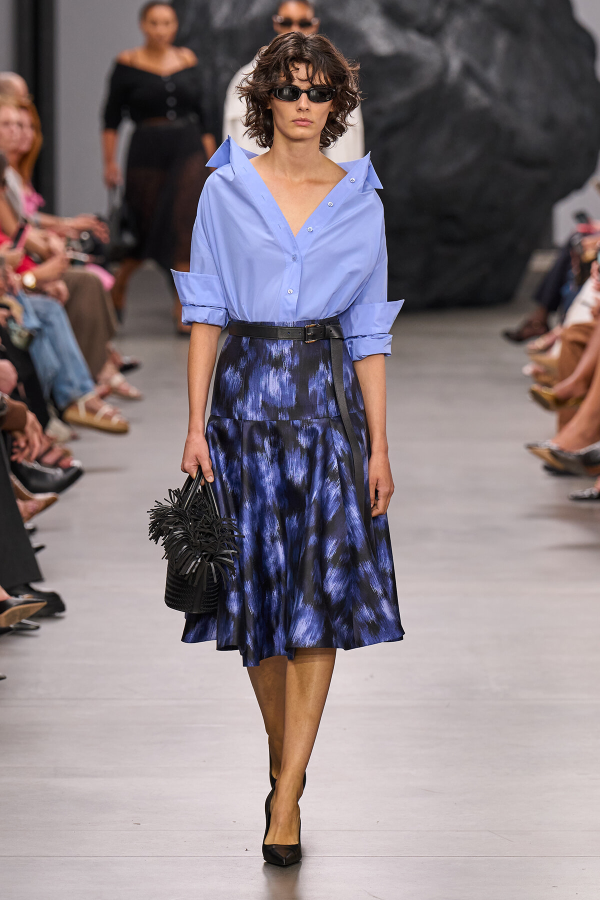 Model walking a runway in a light blue button-down blouse with rolled sleeves, a dark blue printed midi skirt, and a black belt, carrying a black fringe handbag while wearing sunglasses.