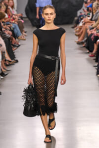 Model walks the runway in a black short-sleeve top and a netted crochet skirt with fringe, cinched by a belt, carrying a spiky black handbag and wearing black slide sandals.