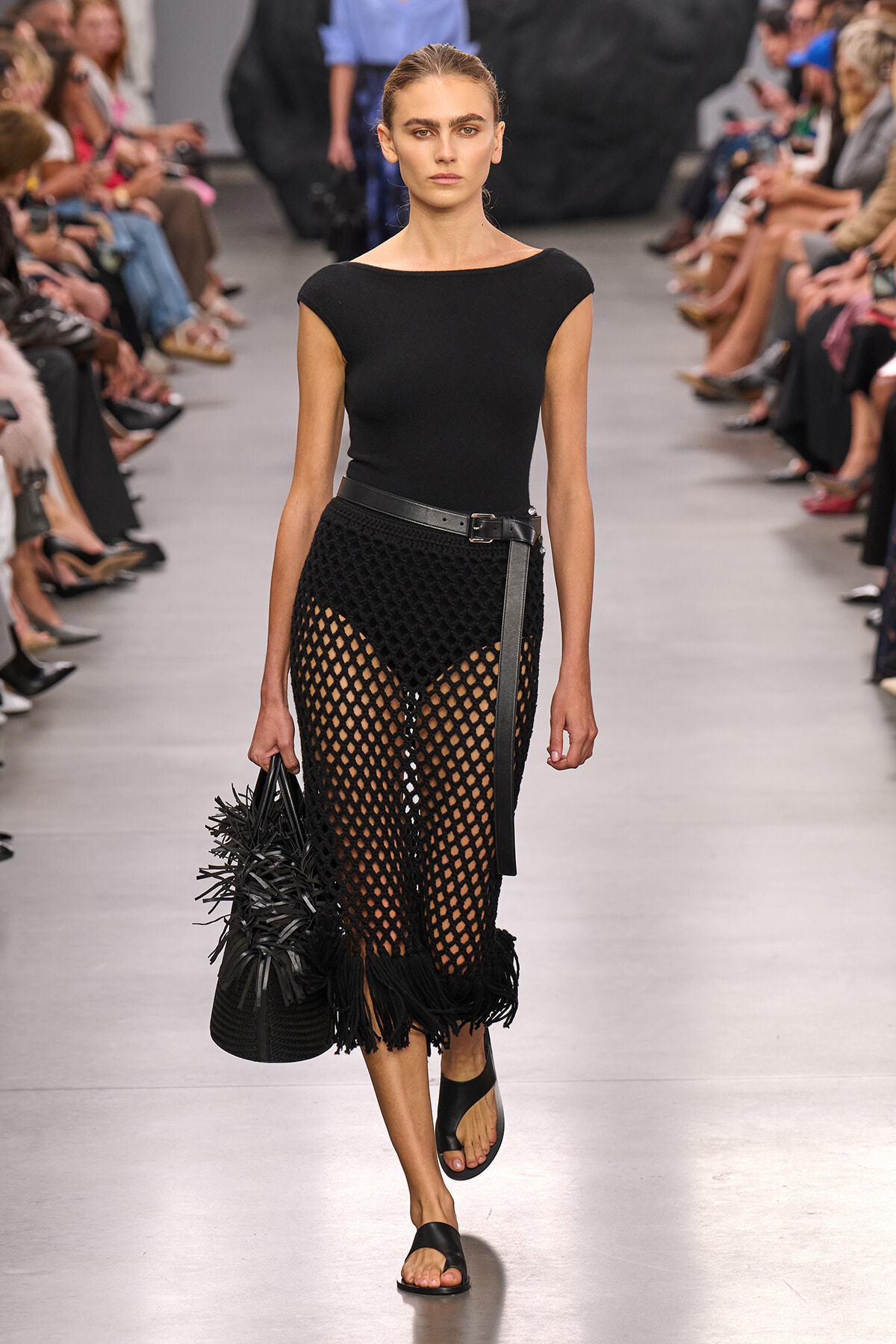 Model walks the runway in a black short-sleeve top and a netted crochet skirt with fringe, cinched by a belt, carrying a spiky black handbag and wearing black slide sandals.