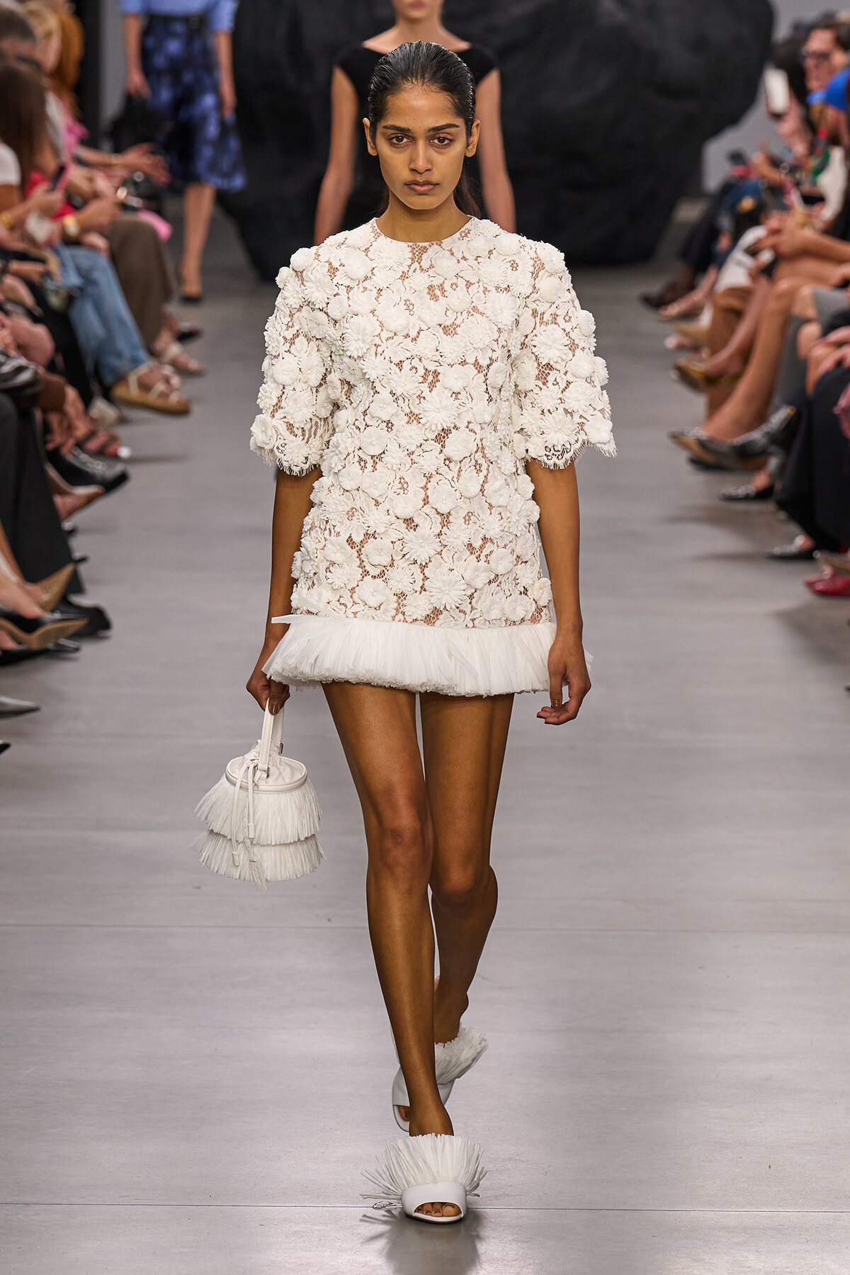 Runway model in a short white lace dress with 3D floral appliqués and a fringed hem, carrying a fringed white handbag.