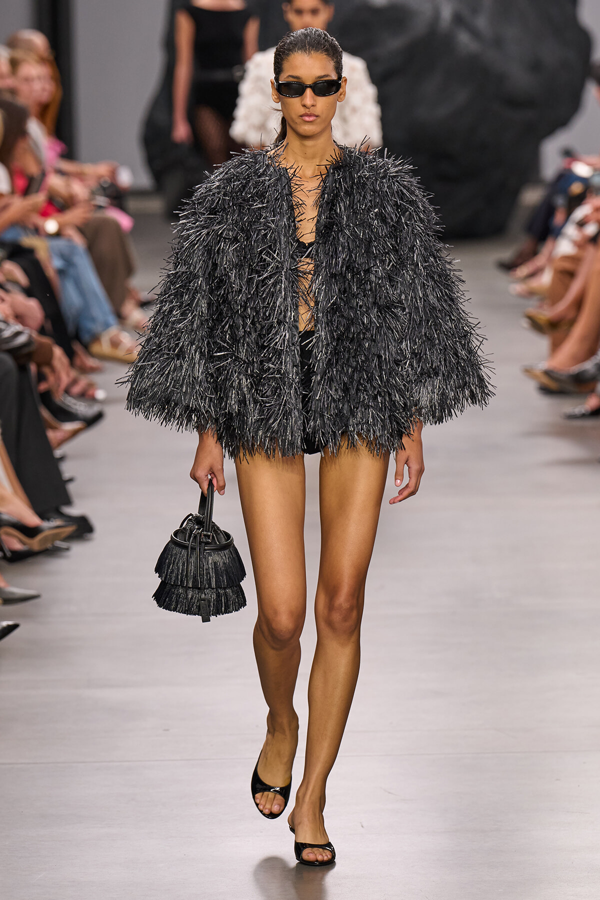 Model on a runway wearing a shaggy black fringe coat, dark sunglasses, and a fringed black bucket bag with open-toe heels.