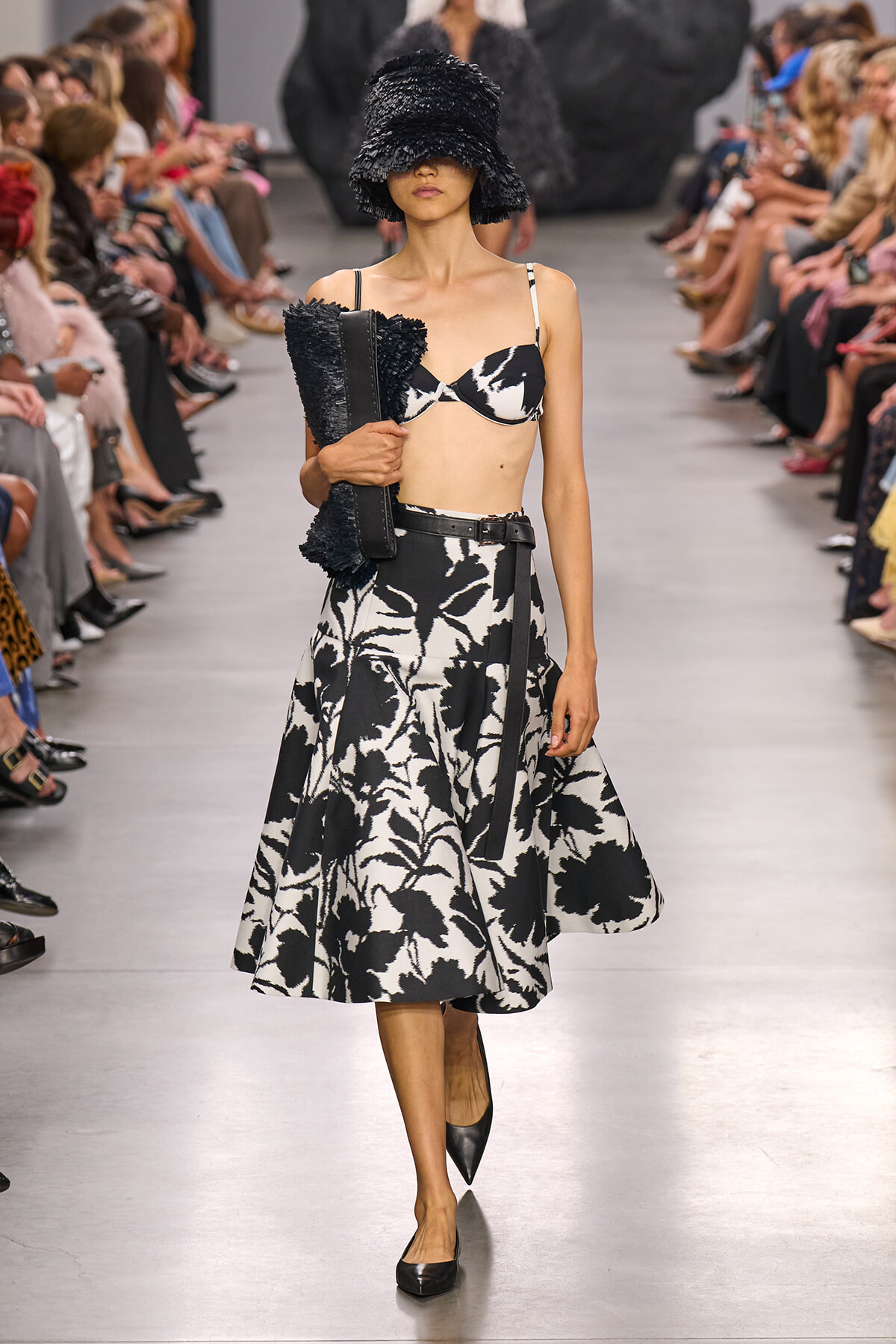 Runway model in a black-and-white floral midi skirt with a matching bikini top, wide textured hat, and clutch.