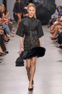 Model on a runway wearing a black blouse with a wide belt, a lace skirt with a feathered fringe hem, black heels, and a tiered black fringe handbag.