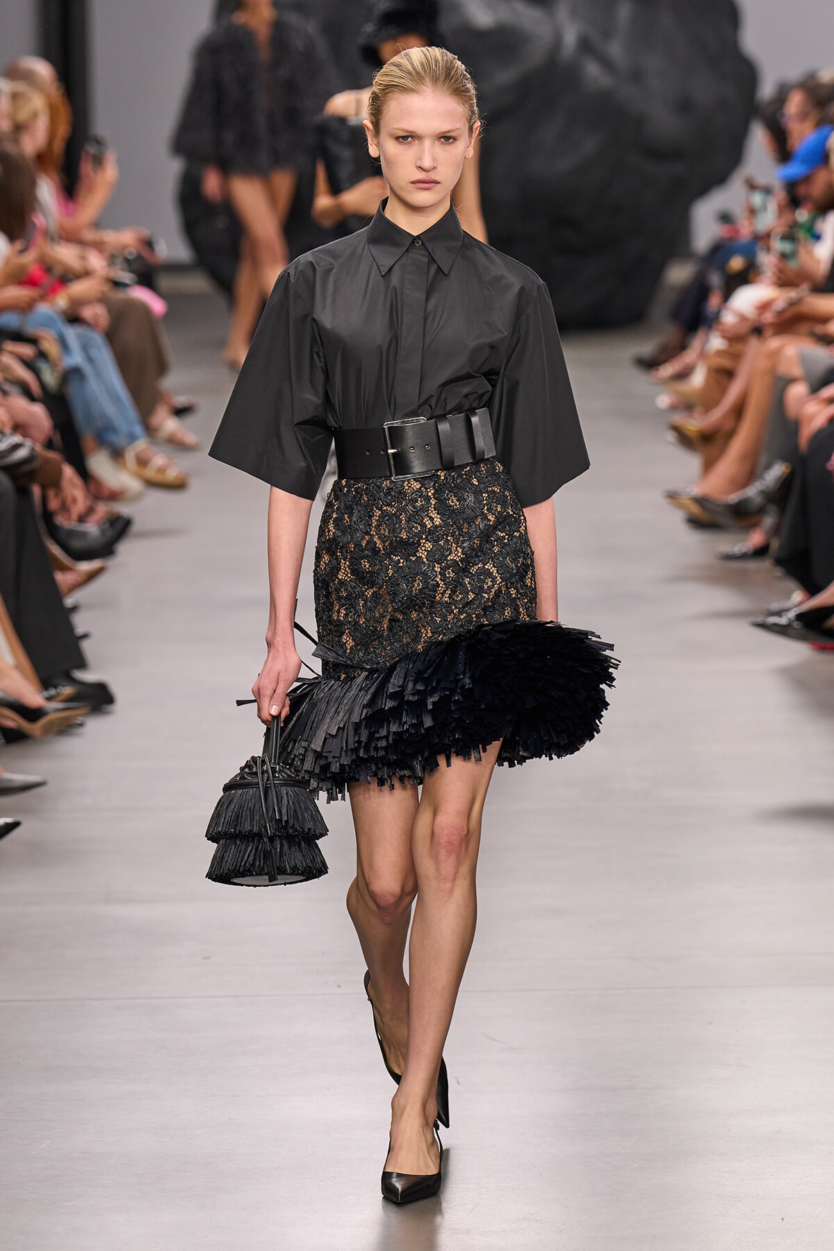 Model on a runway wearing a black blouse with a wide belt, a lace skirt with a feathered fringe hem, black heels, and a tiered black fringe handbag.