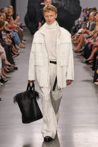 Male model walks a runway in an all-white ensemble: oversized jacket, turtleneck sweater, wide-leg pants, and a large black woven tote bag.
