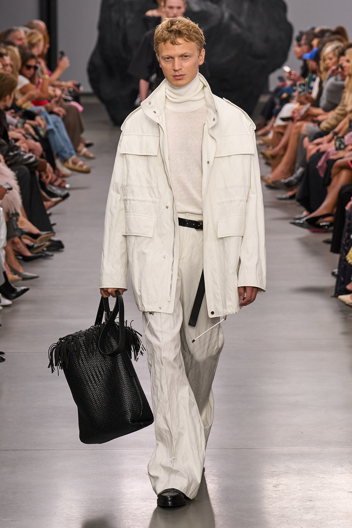 Male model walks a runway in an all-white ensemble: oversized jacket, turtleneck sweater, wide-leg pants, and a large black woven tote bag.