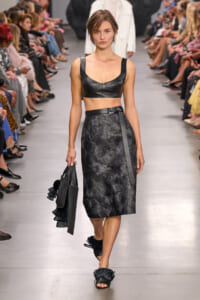 Model on a runway wearing a black leather crop top and a high-waisted, floral-embossed midi skirt, carrying a black fringe handbag.