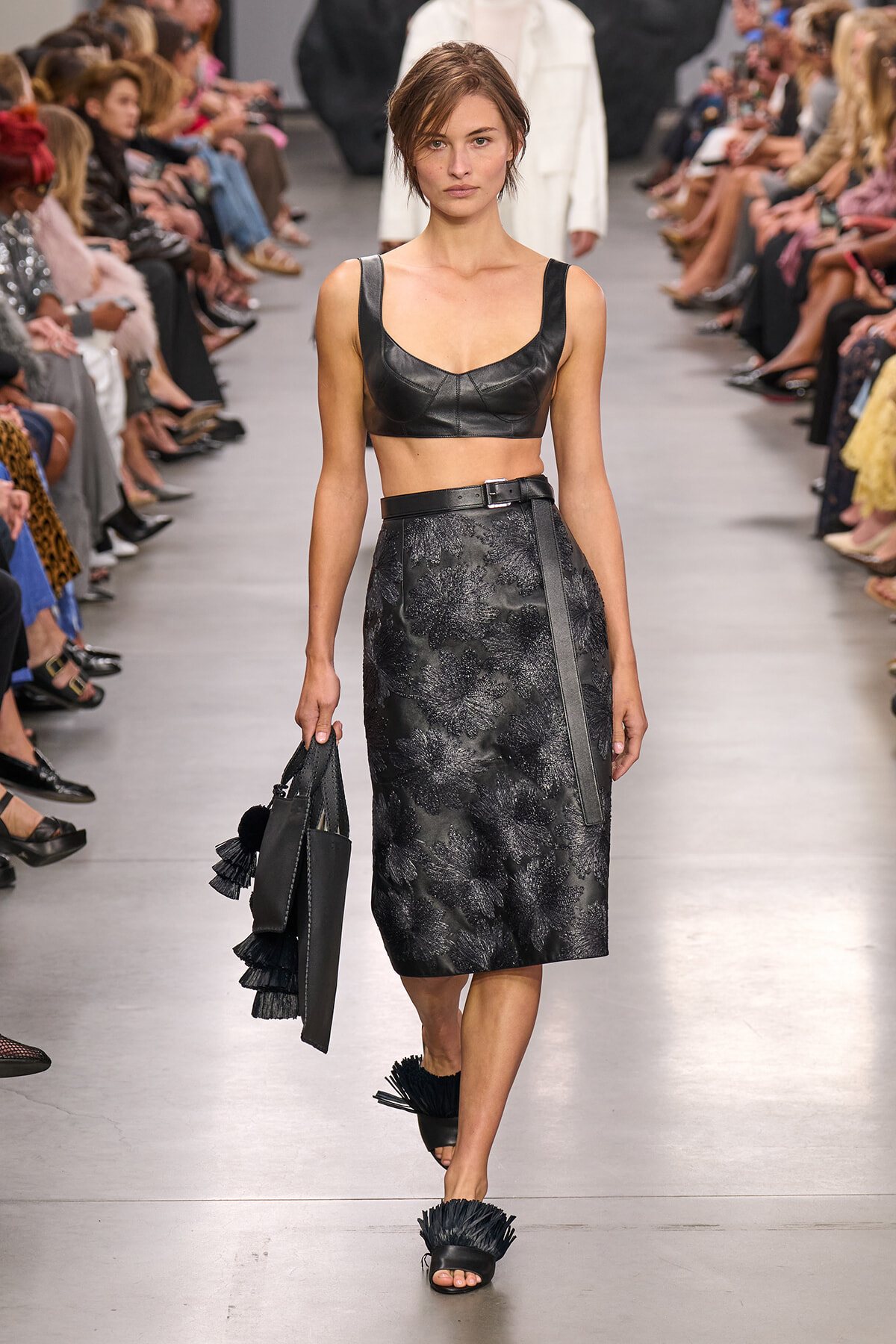 Model on a runway wearing a black leather crop top and a high-waisted, floral-embossed midi skirt, carrying a black fringe handbag.