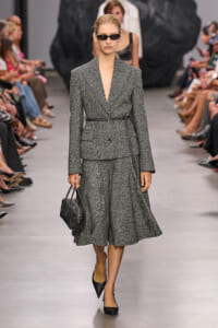 Model on a runway wearing a gray tweed suit with belt and matching midi skirt, carrying a black handbag and wearing black sunglasses.