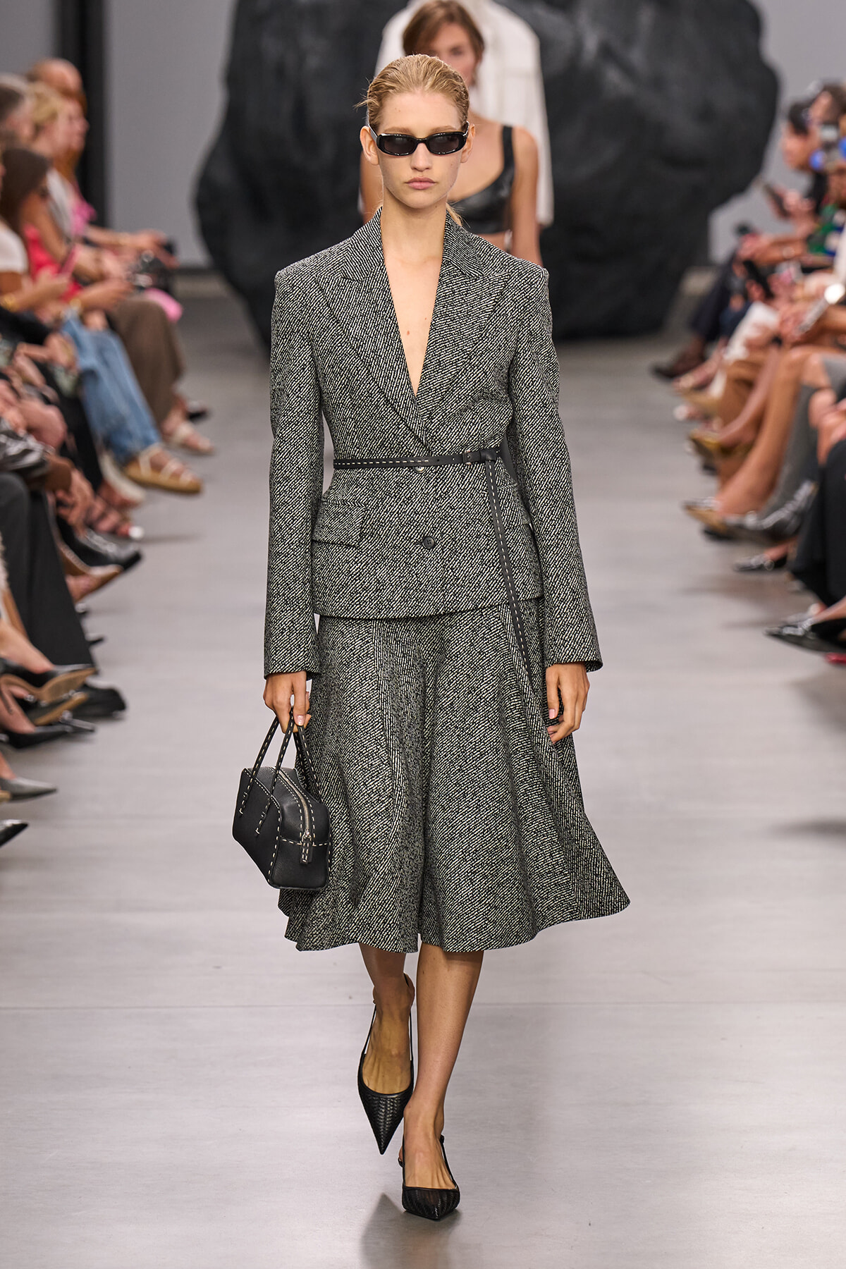Model on a runway wearing a gray tweed suit with belt and matching midi skirt, carrying a black handbag and wearing black sunglasses.
