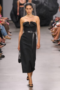 Model walking the runway in a black strapless midi dress with a bow neckline and a thin belt, carrying a small black handbag.