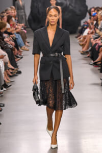 Model walking a runway in a black tailored blazer belted at the waist, paired with a black lace midi skirt and white heels, carrying a small black handbag; fashion show setting with seated audience along the catwalk.
