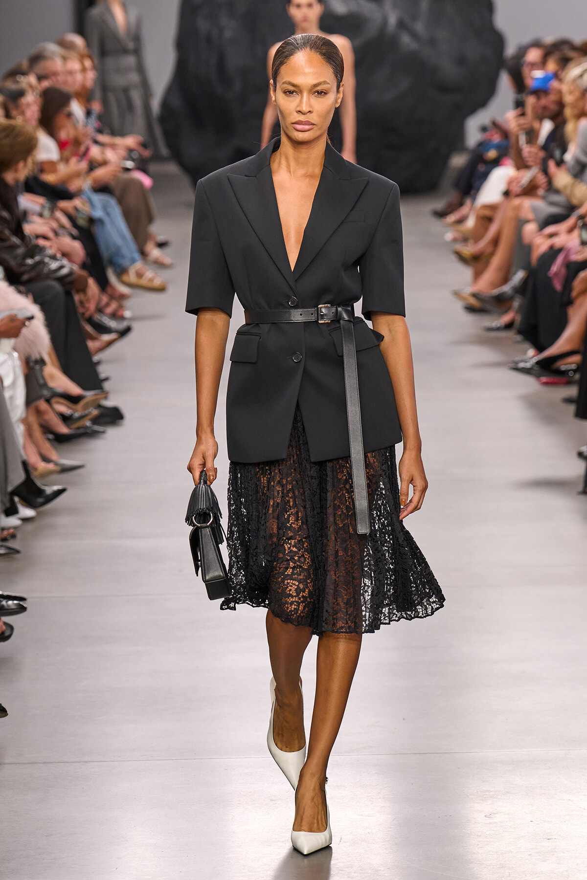 Model walking a runway in a black tailored blazer belted at the waist, paired with a black lace midi skirt and white heels, carrying a small black handbag; fashion show setting with seated audience along the catwalk.