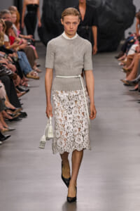Model walking a runway in a light gray knit short-sleeve sweater, a white lace midi skirt, and a white belt, carrying a white handbag.