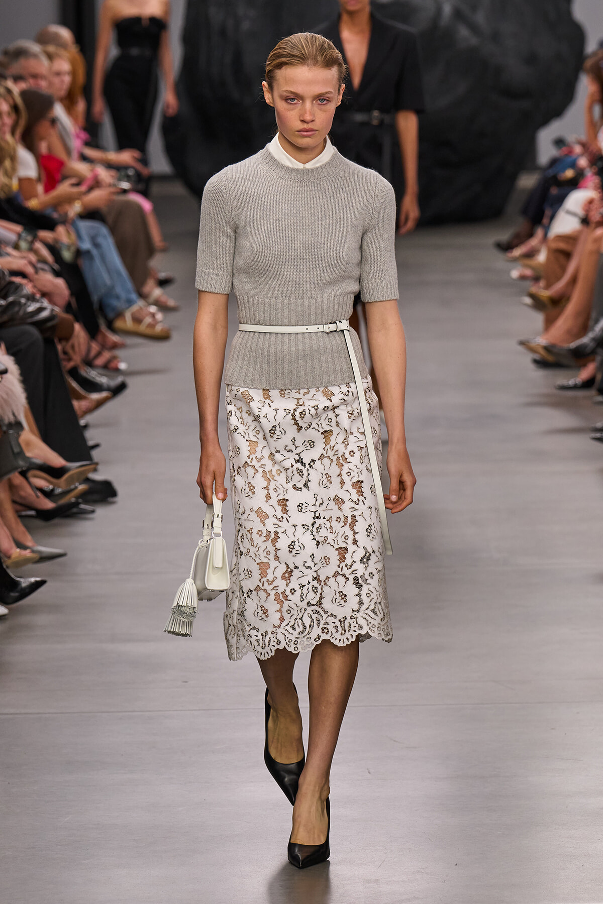 Model walking a runway in a light gray knit short-sleeve sweater, a white lace midi skirt, and a white belt, carrying a white handbag.