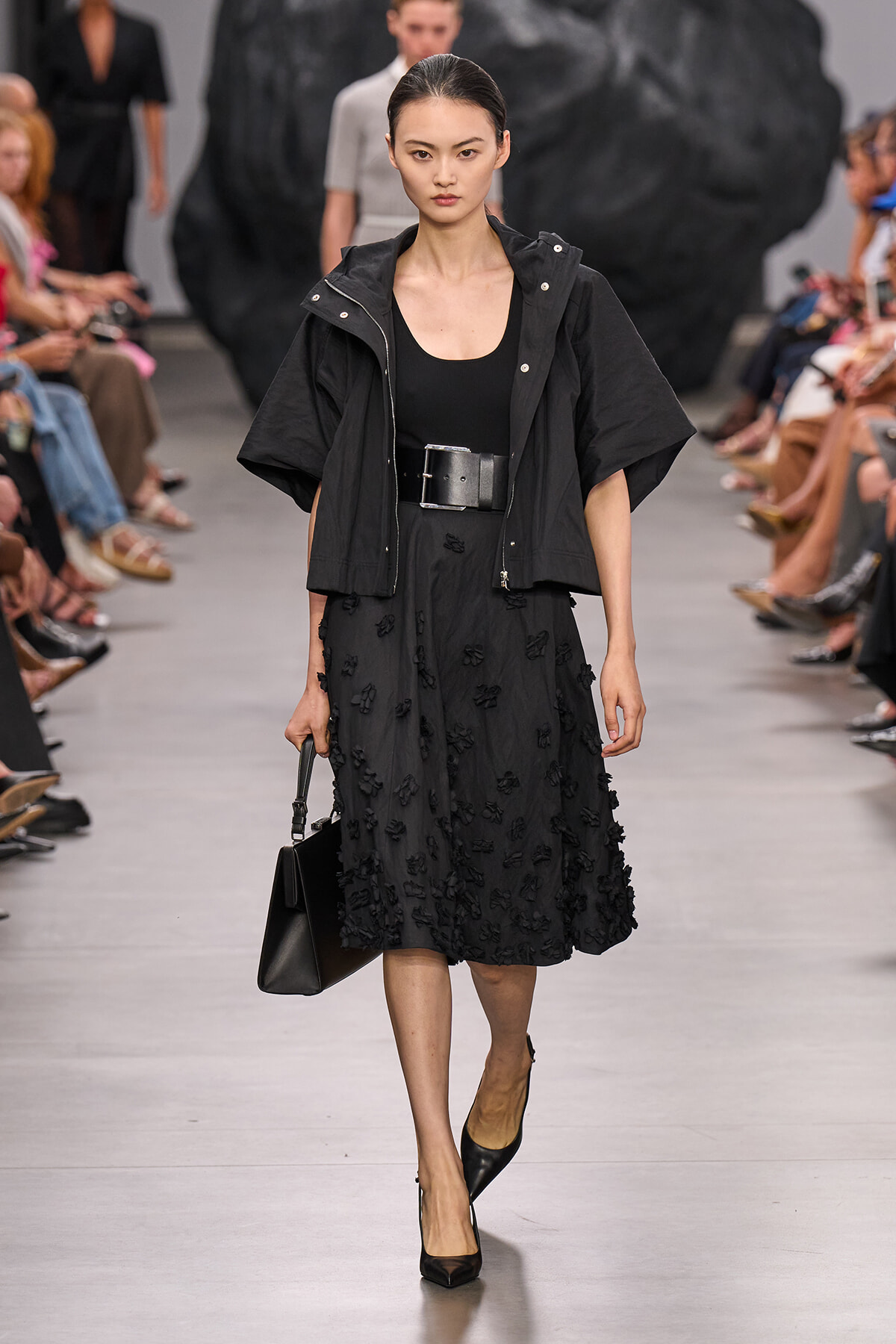 Runway model in an all-black ensemble: cropped jacket, black top, textured midi skirt with appliqués, wide belt, black heels, and a structured black handbag.